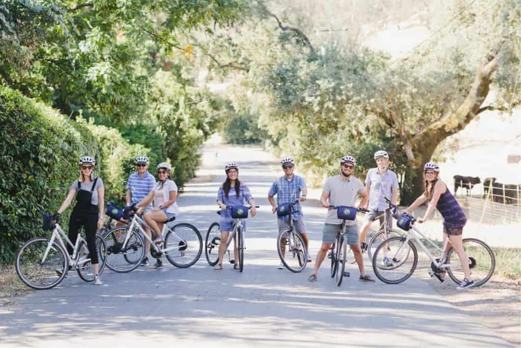 A group of eight people riding bikes pause for a photo along a shaded country road