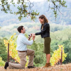 A man kneeling on one knee holds a small open box and looks up at his girlfriend, who is covering her mouth with vineyards in the background.