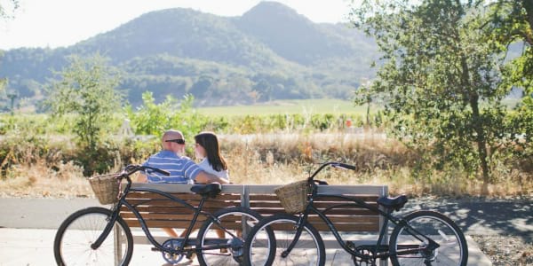 A couple sits close on a bench in front of the vineyards with cruiser bikes parked behind them.