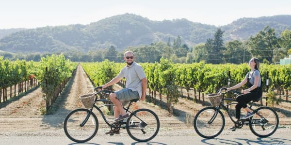 A man and woman ride cruiser bikes in front of rows of vineyards