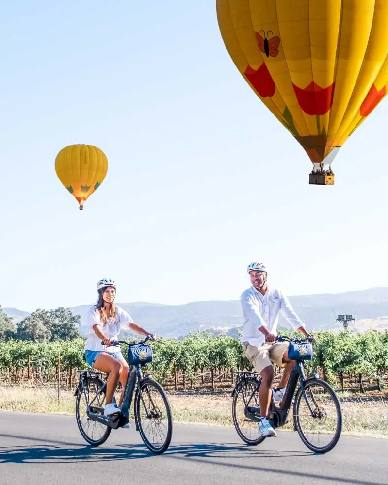 A couple rides electric bikes past the vineyards with hot-air balloons in the background