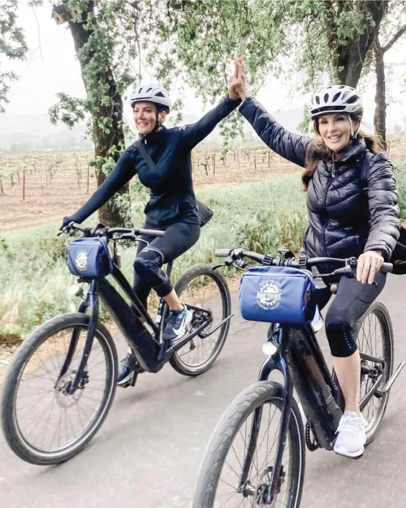 Two women ride electric bikes side by side while giving each other a high-five