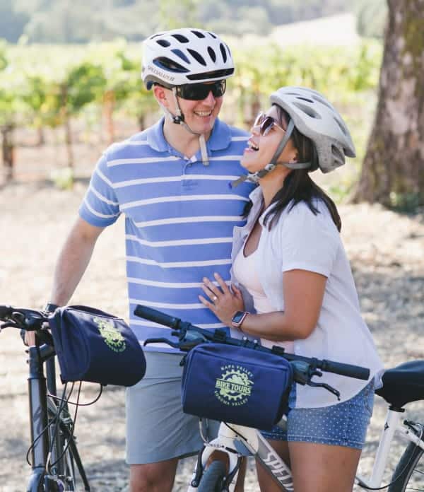 A couple wearing helmets pauses on the side of the road with their bikes and shares a laugh.