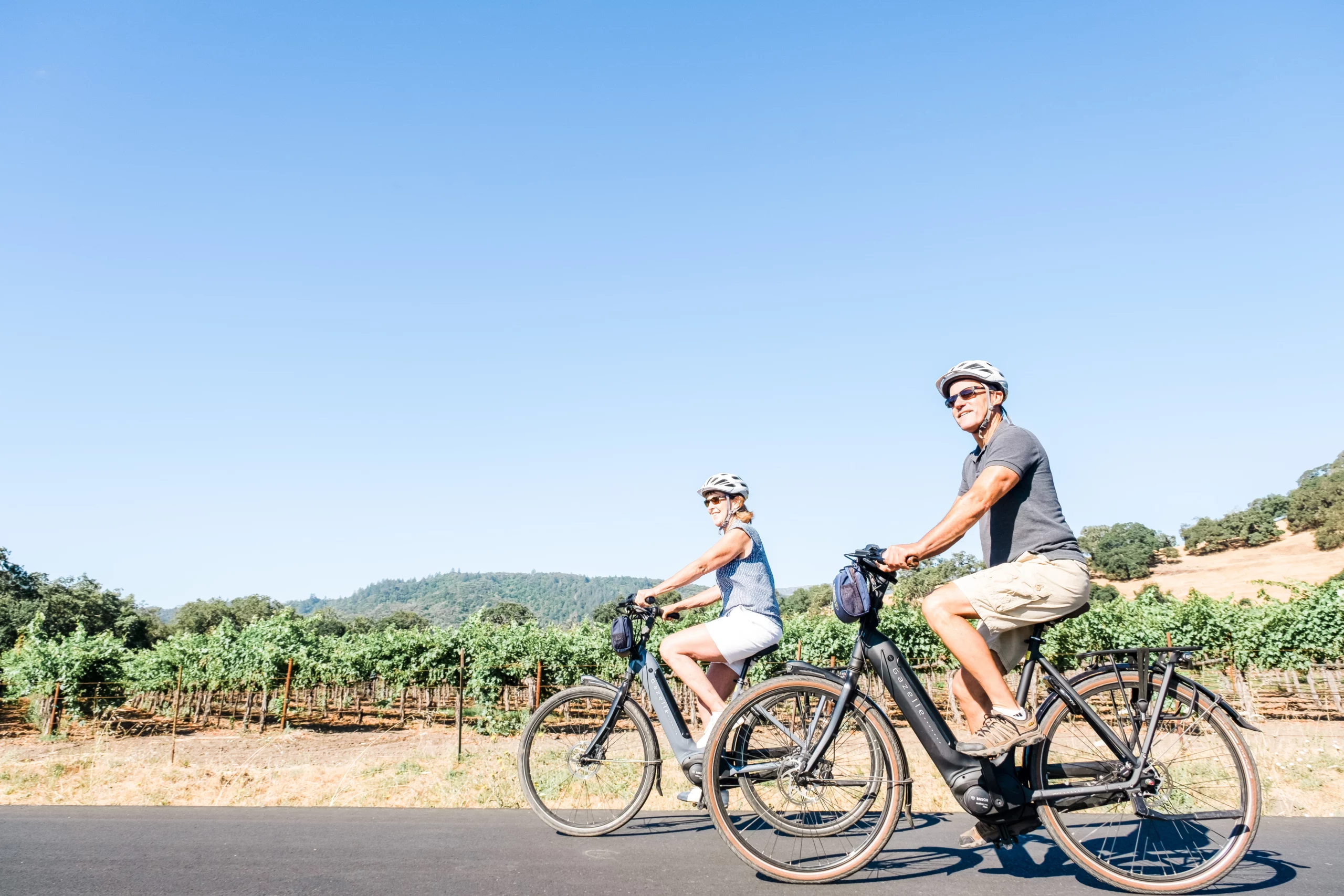 A couple rides electric bikes along a road bordered by vineyards.
