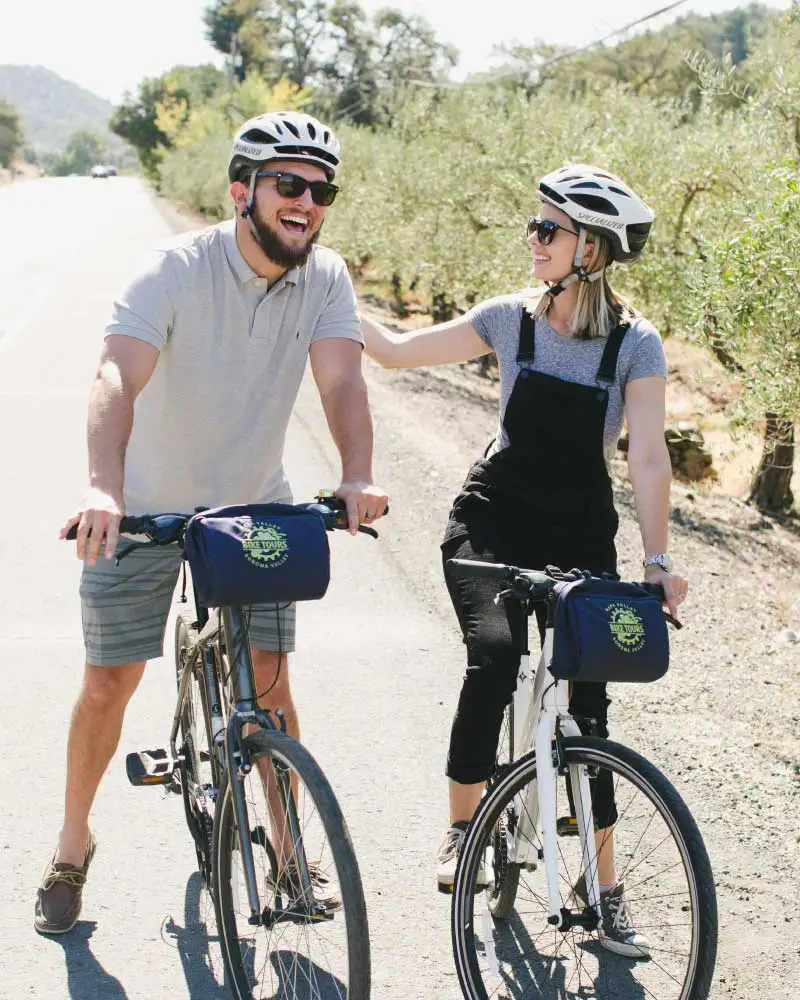 A couple riding bikes laughs and smiles while stopped along a road bordered by olive trees.