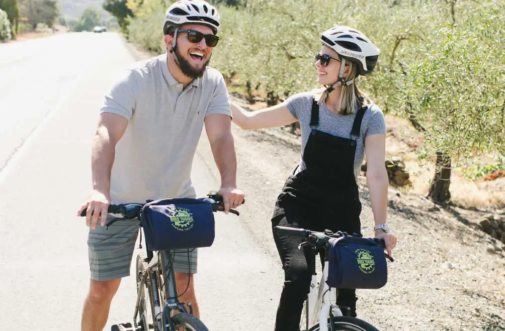 A couple on bikes pauses along the road in front of olive trees.