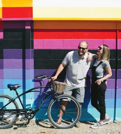 A couple laughs and poses with a cruiser style bike in front of a colorful mural.