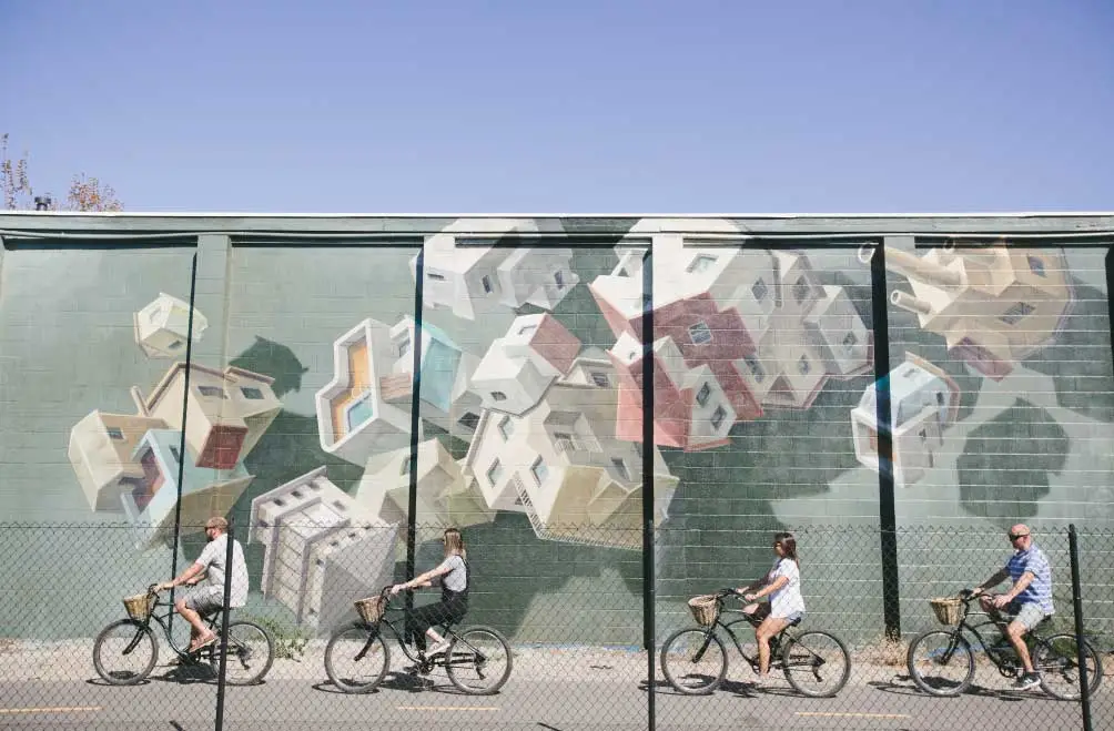 Four people ride beach cruiser style bikes past a colorful mural depicting tumbling houses