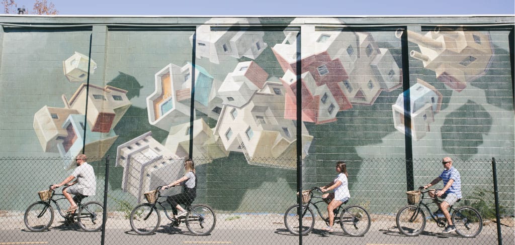 Four adults ride bikes single file past a mural depicting houses tumbling from the sky