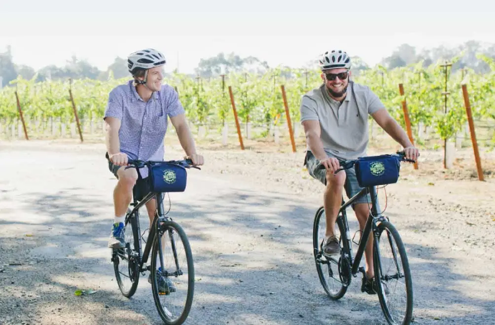 Two men smile as they ride bikes side by side along a quiet country road.