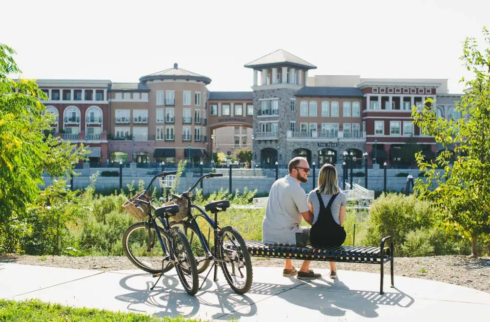 A couple sits on a bench overlooking the Napa riverfront with cruiser style bikes parked beside them.