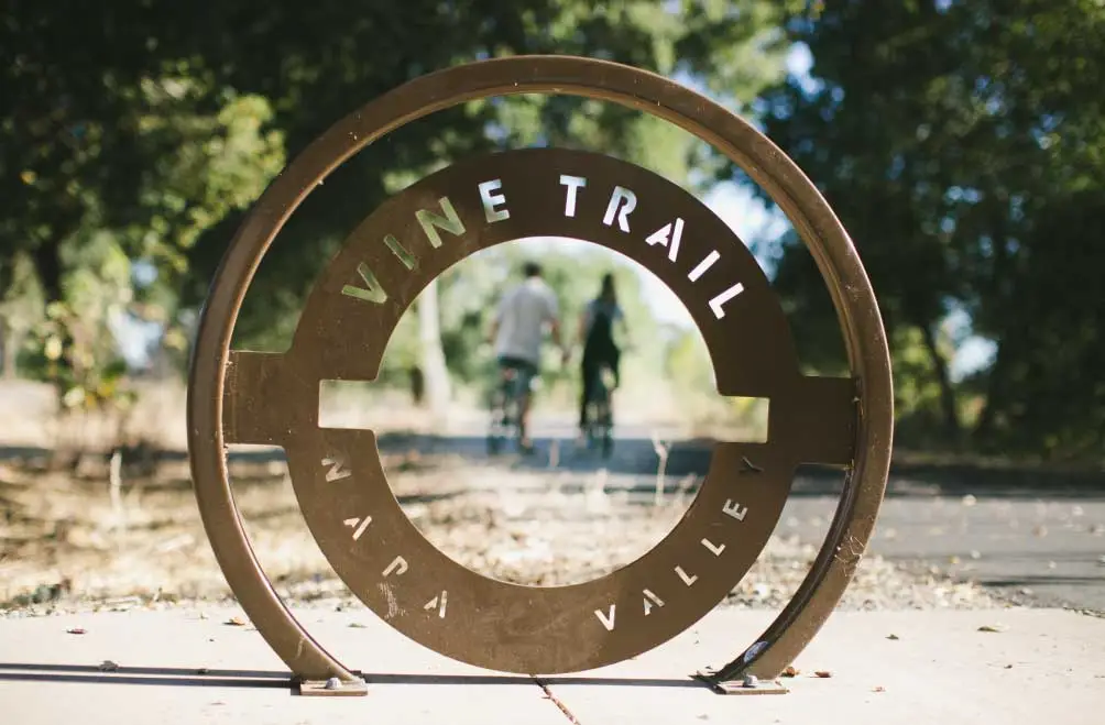 A couple riding bikes in the distance is framed by a circular Napa Valley Vine Trail sign in the foreground.